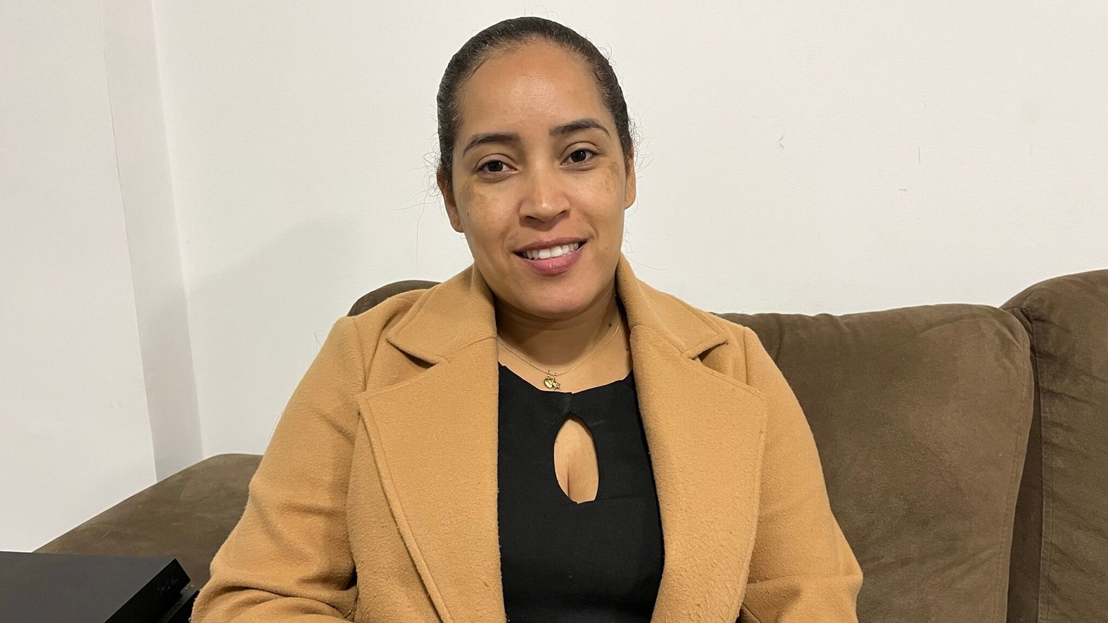 Amanda sorri para a foto sentada em um sofá. Ela usa blusa preta e casaco marrom